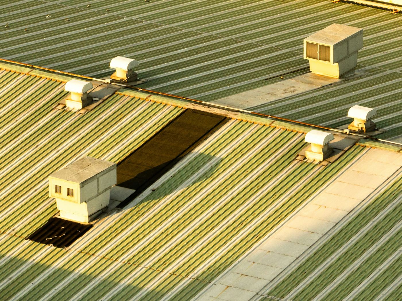 Oblique aerial view of a corrugated metal factory roof with rooftop ventilation units and two dark replacement panels in low sun