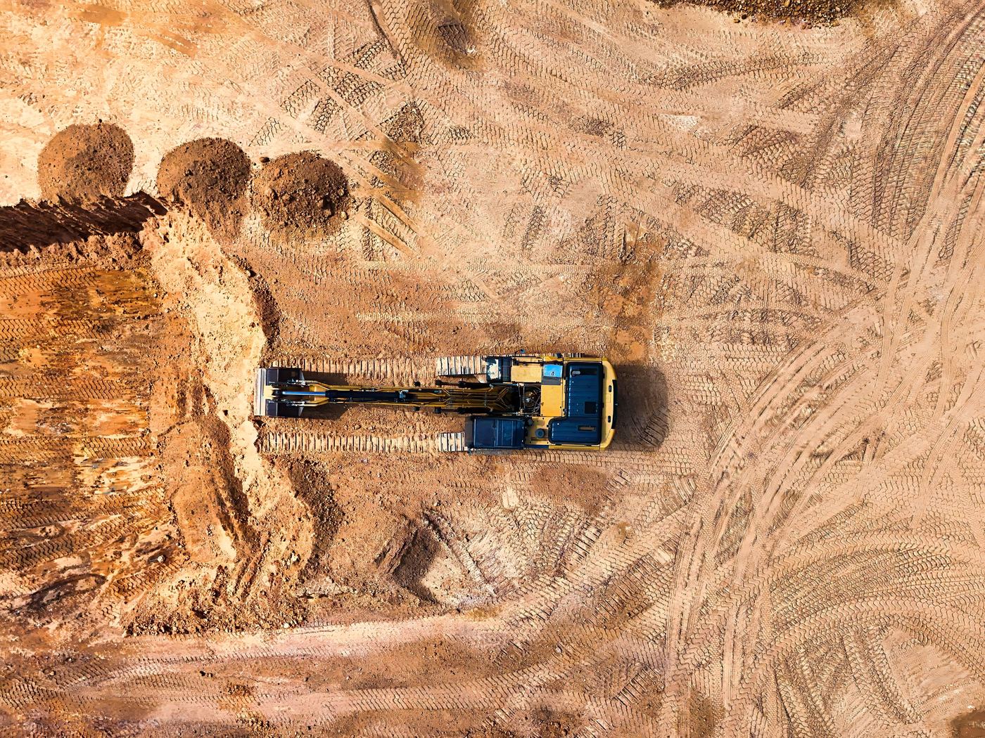 Drone view straight down on a tracked excavator cutting a bench on a cleared earthworks pad, with spoil heaps and plant tracks in sandy ground