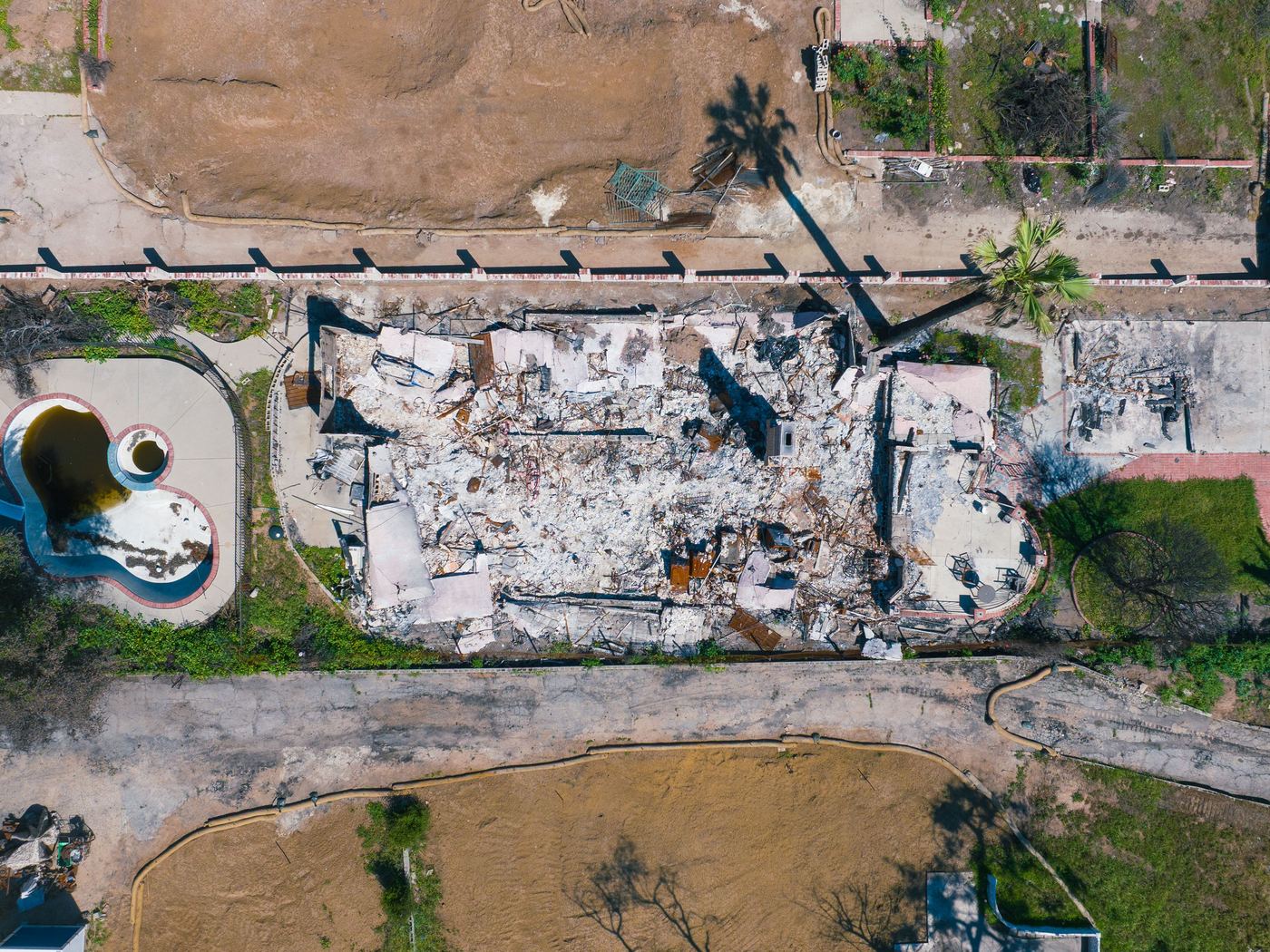 Straight-down aerial of a fire-destroyed house: collapsed walls and rubble inside the footprint, an intact pool beside it and scorched ground around