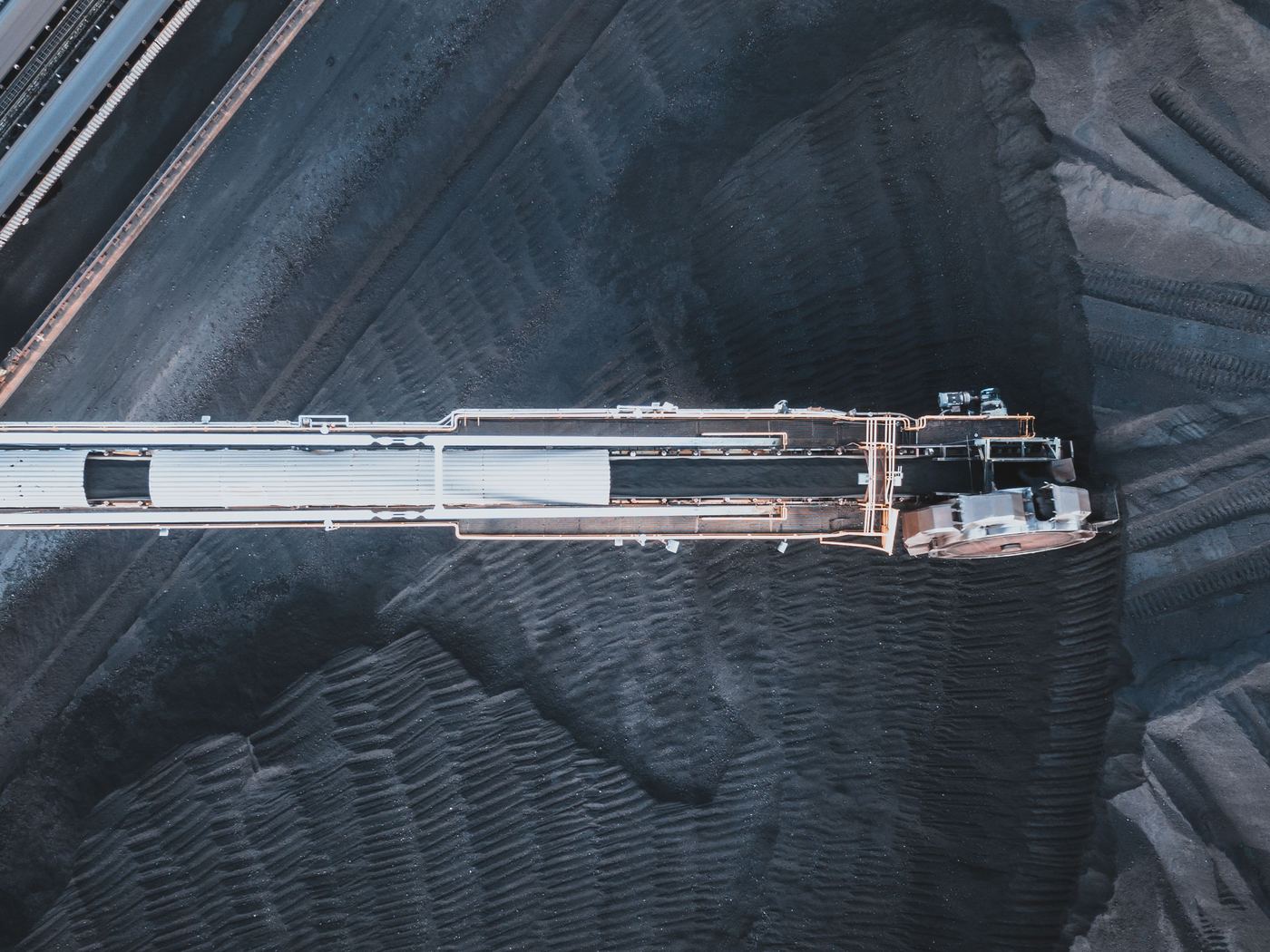 Top-down aerial of a white stacker-reclaimer boom conveyor across a dark ridged coal stockpile, with rail lines at top left