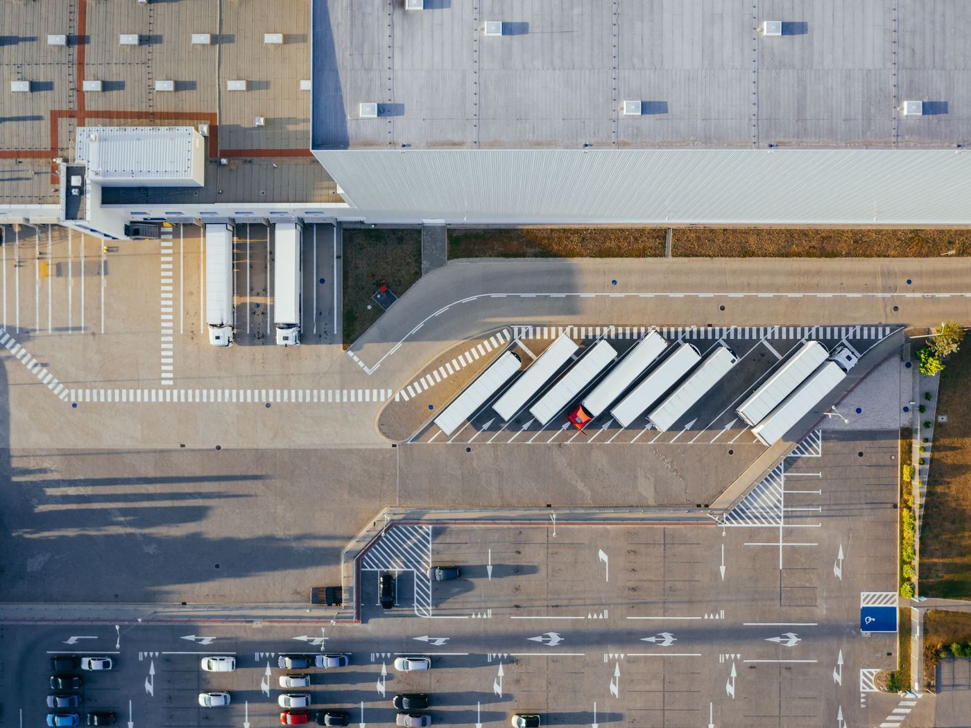 Top-down drone view of a logistics warehouse roof with skylights, loading docks, parked semi-trailers and a line-marked car park.
