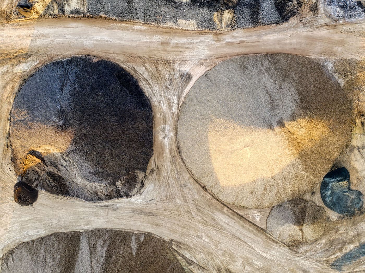 Straight-down aerial of a gravel quarry: two large circular stockpiles, one dark and one light, divided by pale haul roads.